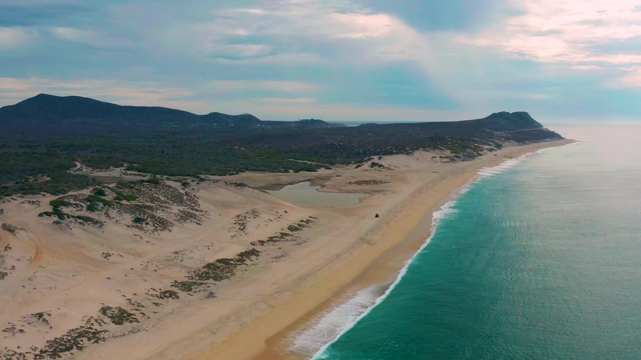 Aerial view over Laguna Beach, Baja California Sur with sandy dunes in the foreground, turquoise ocean on the right, low mountains inland, and no visible people.