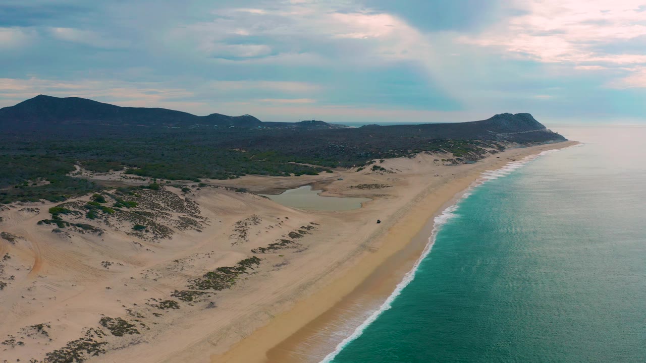 Aerial frame of Laguna Beach, Baja California Sur with turquoise water beside a pale sandy shore, dune patches inland, dark hills across the horizon, and no visible people.