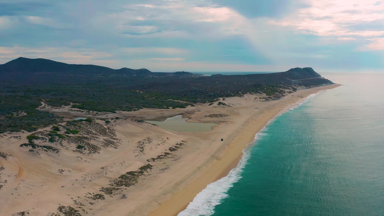 Wide aerial frame of Laguna Beach, Baja California Sur, showing the shoreline angling toward a rocky headland, with dunes and scrub inland and no visible people.