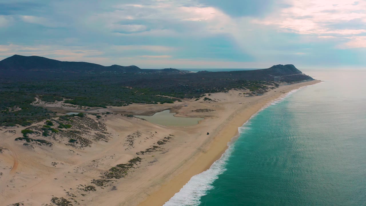 Aerial view of Laguna Beach in Baja California Sur with more open water filling the right side, a continuous sand strip, dune patches, inland vegetation, hills, and no visible people.