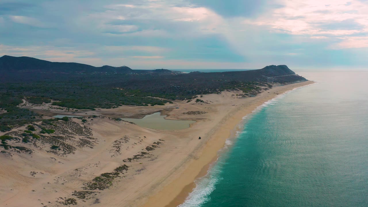 Wide aerial frame of Laguna Beach, Baja California Sur, showing a diagonal surf line, pale dunes, scrubland, dark inland hills, a headland in the distance, and no visible people.
