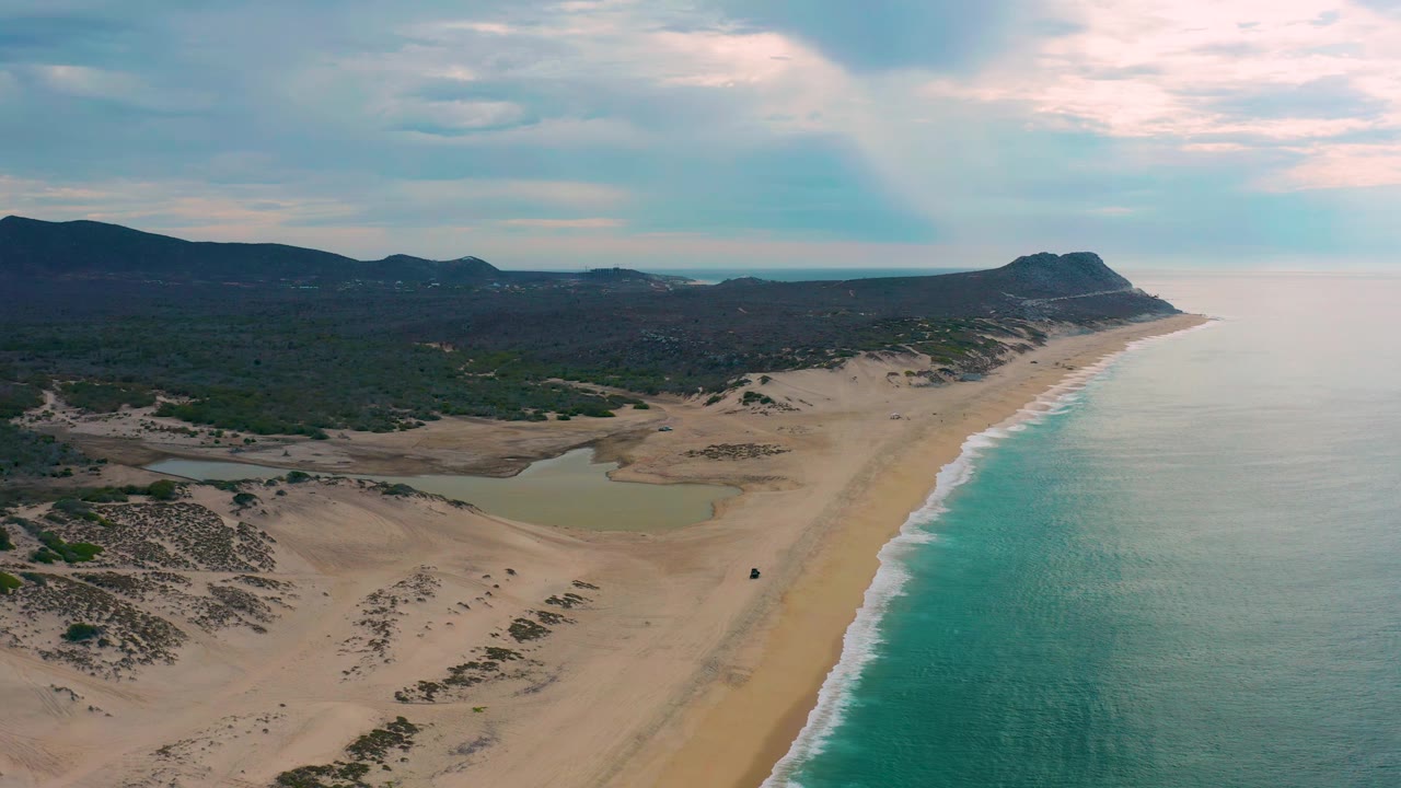 High aerial frame of Laguna Beach, Baja California Sur, showing the shoreline angled into the distance, lagoon water left of the beach, low hills beyond, and no visible people.