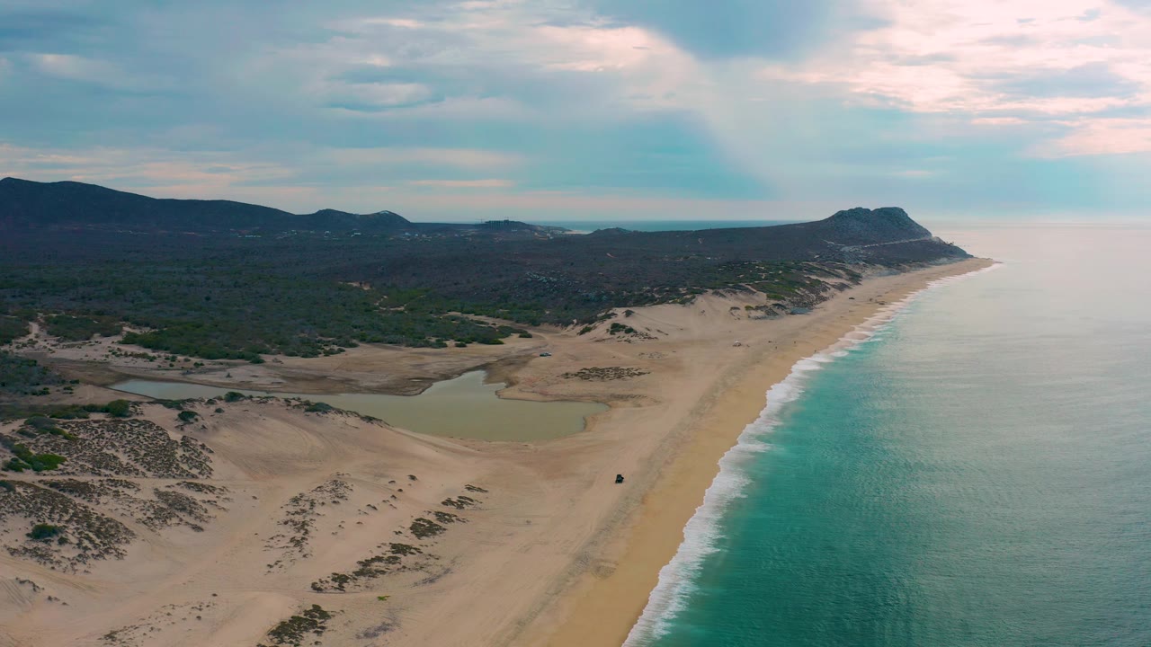 Drone view along Laguna Beach, Baja California Sur, with turquoise water on the right, broad sand and dune scrub on the left, distant mountains ahead, and no visible people.