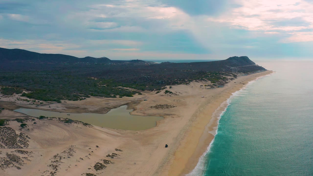 Drone view along beach with lagoon, surf, dunes, vegetation, and distant headland