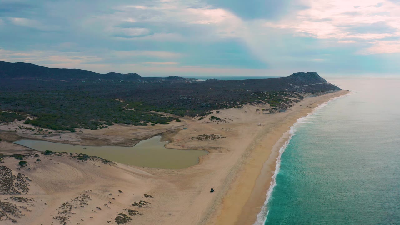 Laguna Beach, Baja California Sur appears from above with the ocean filling the right side, pale waves tracing the beach, a lagoon and dune field in the lower left, mountain ridges across the horizon, and no visible people.