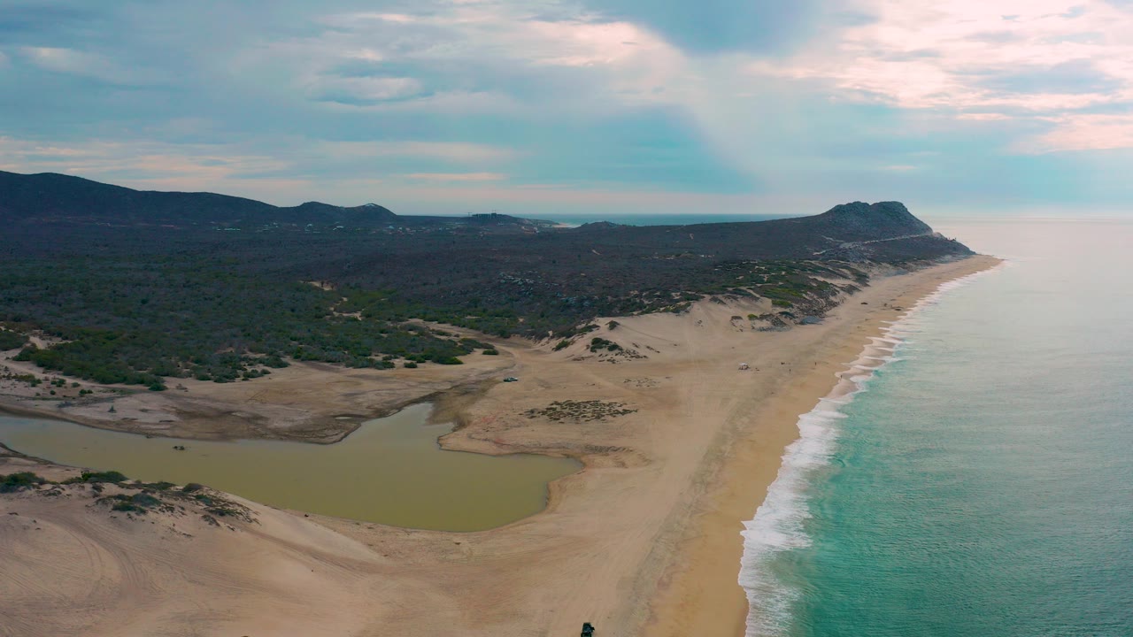 Aerial frame over Laguna Beach BCS, Mexico, with a green lagoon in the lower left, sandy dunes, turquoise surf along the right edge, distant hills, and no visible people.