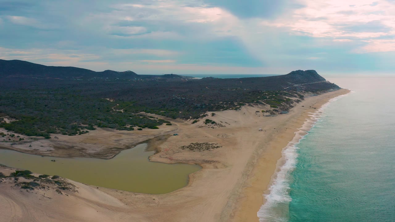 Drone frame over Laguna Beach BCS with the lagoon clipped at the lower left, wider sand across the center, surf running up the right side, inland scrub, hills, and no visible people.