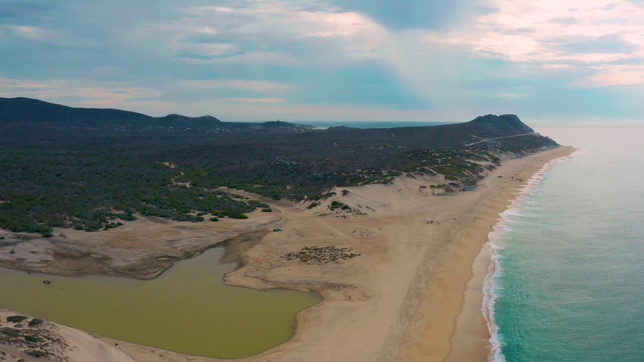 Wide aerial coastline with lagoon, beach, surf, dark vegetation, hills, and cloud cover.