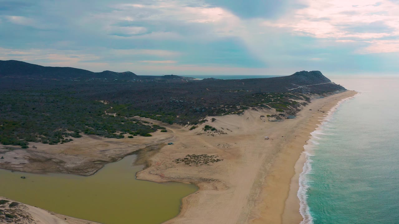 Drone-style aerial view over Laguna Beach, Baja California Sur, with turquoise water and white surf on the right, sand and lagoon below, dark scrubland inland, low mountains on the horizon, and no visible people.