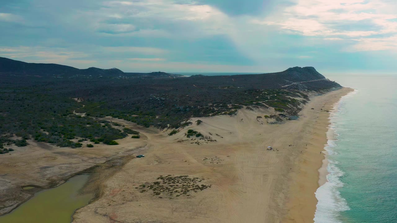 Drone frame over Laguna Beach showing the shoreline stretching into the distance, surf breaking along the right edge, darker scrubland inland, scattered dunes, and no visible people.
