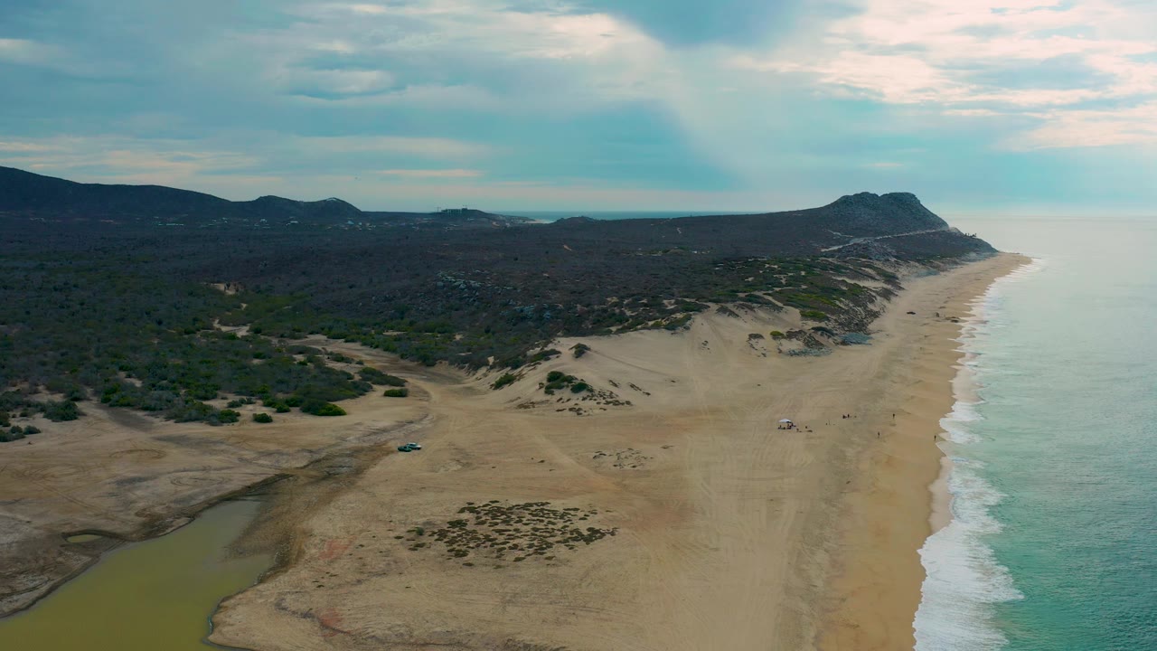 High aerial frame of Laguna Beach in Baja California Sur with teal water to the right, a broad sand strip, dune patches near the center, inland hills on the left, and no visible people.