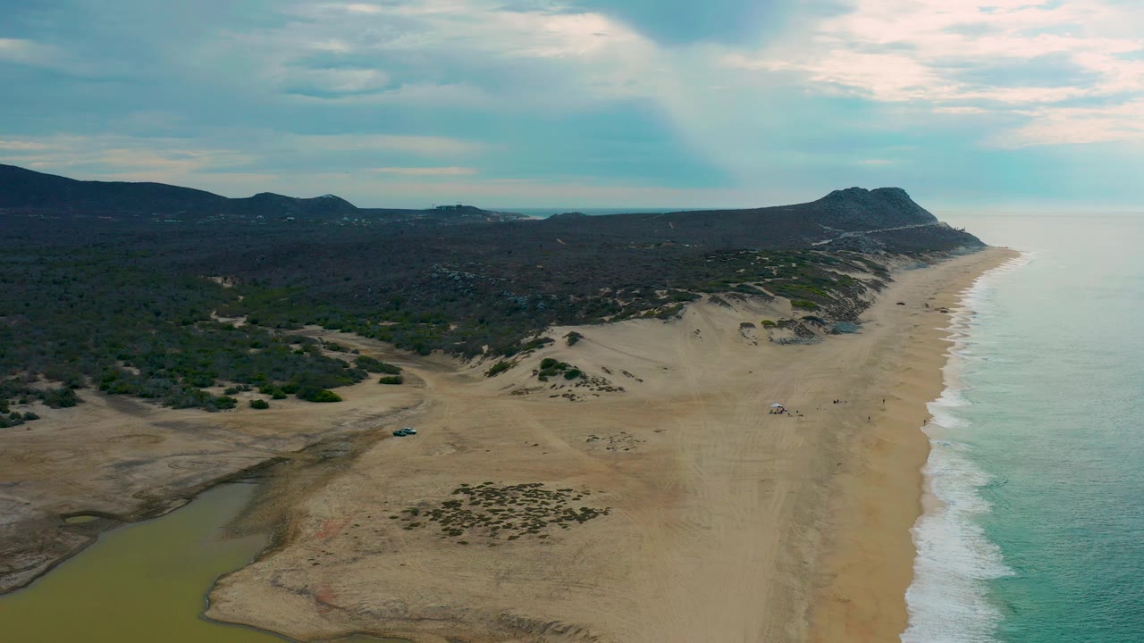 Aerial view of Laguna Beach with a lagoon-like green water area in the lower left, open sand across the middle, ocean surf on the right, dark hills beyond, and no visible people.