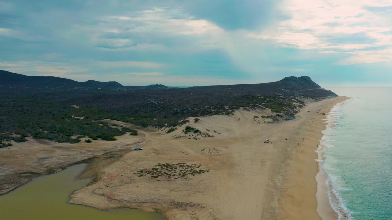 Aerial view of Laguna Beach, Baja California Sur with greenish lagoon water in the foreground, a broad sandy beach, surf on the right, scrubland inland, low hills beyond, and tiny distant beach activity.