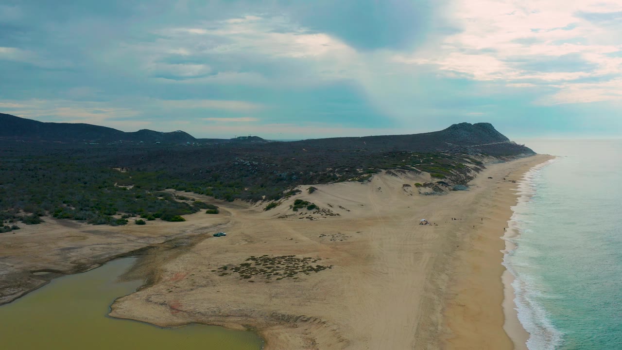 High oblique view of Laguna Beach, Baja California Sur with the lagoon occupying the lower left, a wide pale sand strip, small distant figures or beach items near mid-beach, surf to the right, and dark hills across the horizon.