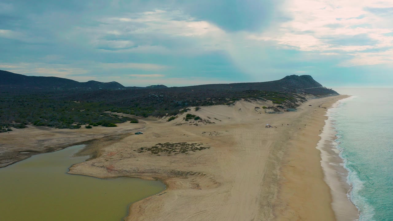 Opening aerial frame of Laguna Beach in Baja California Sur, Mexico, with a green-brown lagoon at lower left, a wide sand strip, ocean on the right, distant hills, and no visible people.