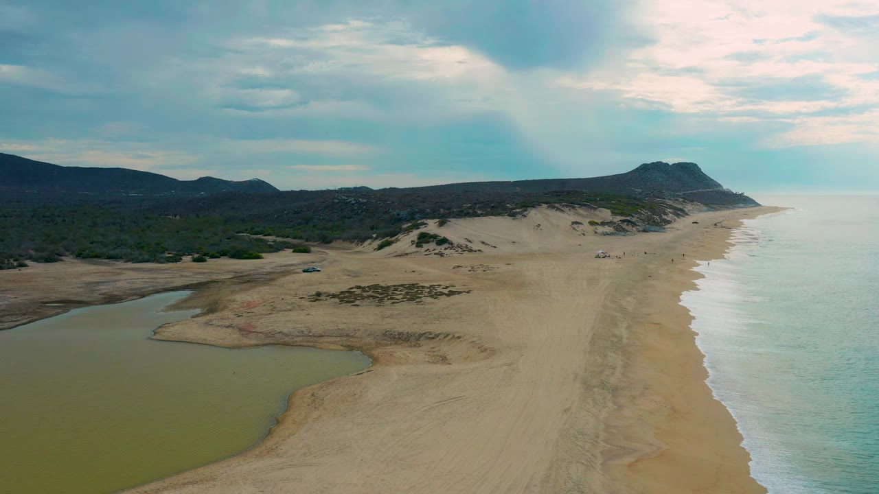 Aerial view of Laguna Beach, Baja California Sur, showing the lagoon water larger in the lower left, pale surf tracing the right shoreline, dunes and scrub inland, distant hills, and no visible people.