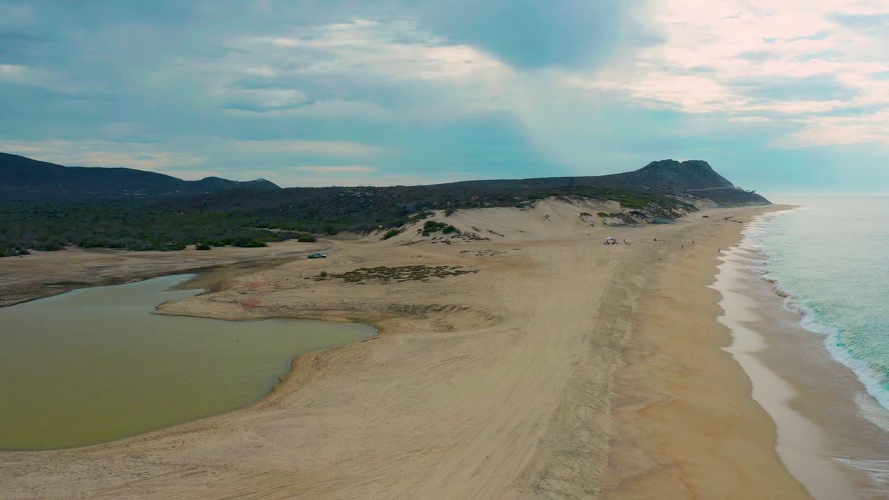 Aerial view of sandy coast with lagoon, surf, dunes, hills, and clouds.