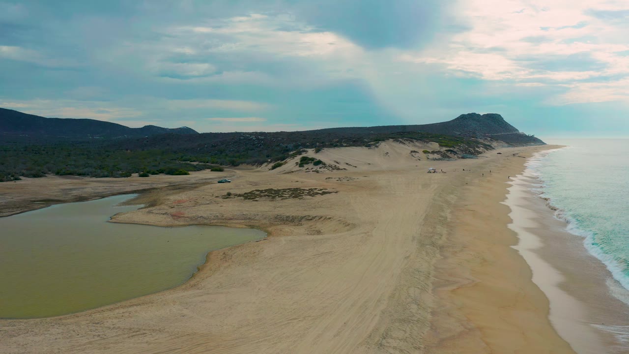 Aerial view of Laguna Beach, Baja California Sur, with the lagoon curving into the sand at left, the ocean wash tracing the right side, distant hills, and no visible people.