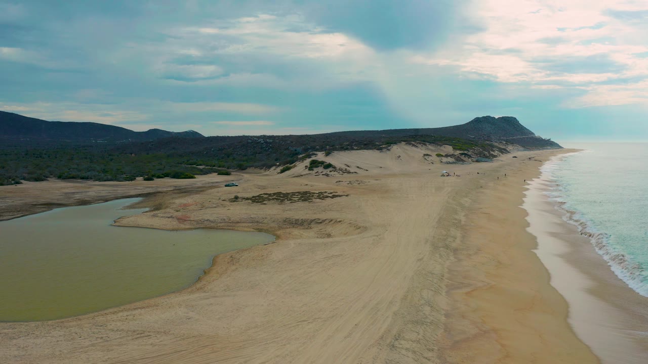 Laguna Beach aerial frame showing the lagoon low on the left, a broad sand corridor through the center, pale waves on the right, a dark distant headland, and no visible people.