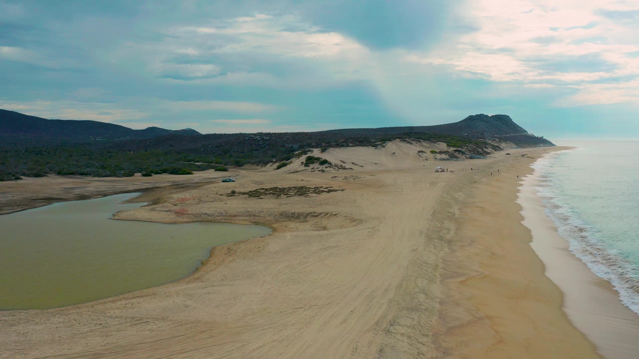 Aerial frame over Laguna Beach, Baja California Sur, where the green lagoon sits in the lower left, the wave wash runs along the right, dunes fill the middle distance, and no visible people appear.