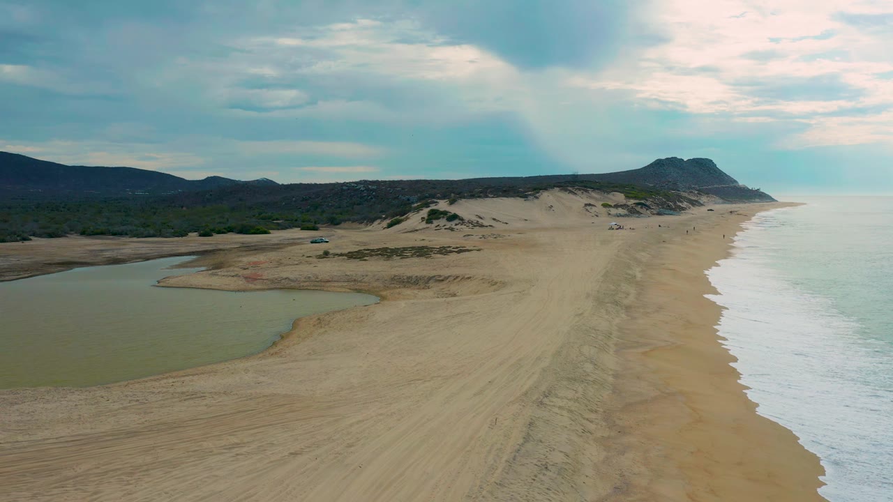 Aerial frame over Laguna Beach, Baja California Sur, Mexico, with the lagoon widening in the left foreground, tire-like tracks running down the sand, ocean foam along the right edge, and no visible people.