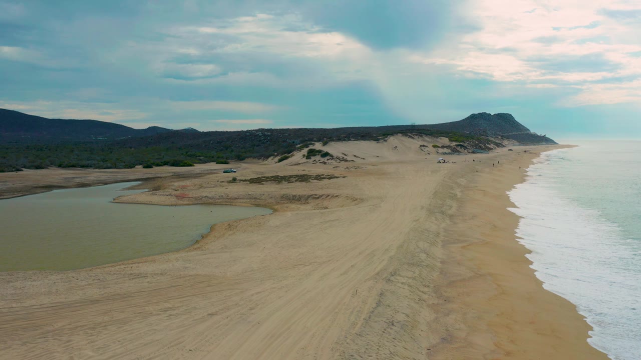 Drone-height view of Laguna Beach in Baja California Sur, Mexico, showing the lagoon tucked into the left side, broad tracked sand through the middle, a narrow surf line on the right, and no visible people.