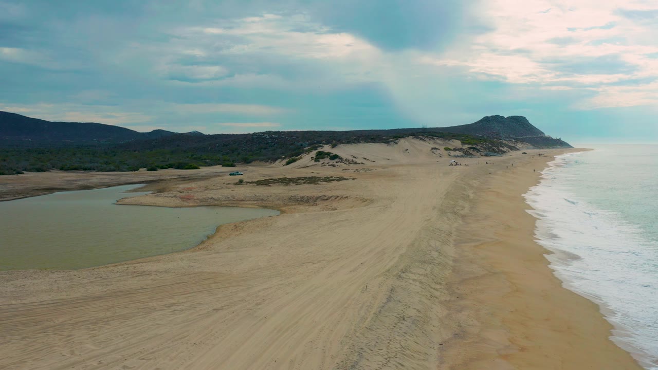 Aerial view over Laguna Beach, Baja California Sur, Mexico, with the lagoon in the lower left, long sand tracks centered, white waves breaking along the right side, darker hills behind the dunes, and no visible people.