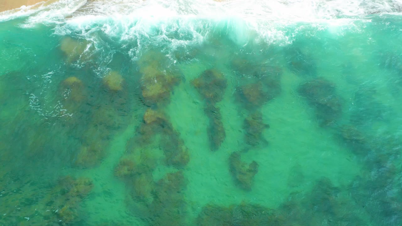 Overhead view at Laguna Beach, Baja California Sur, showing turquoise shallow water, foamy surf along the top edge, dark submerged rocks, and no visible people.