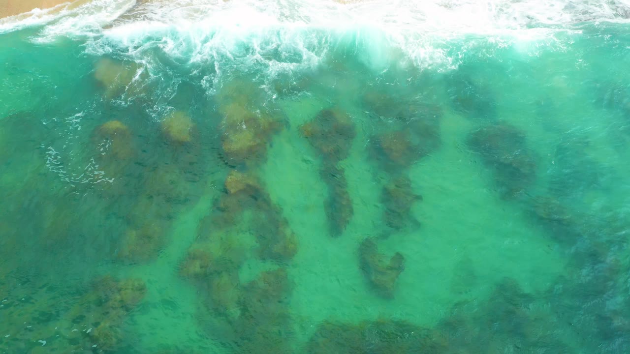 Aerial surf line over green-blue water and underwater rocks.