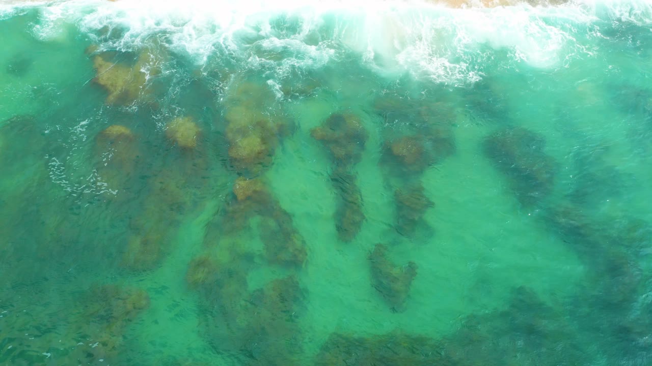 Top-down view at Laguna Beach, Baja California Sur, where teal water covers elongated dark rock shapes and a band of breaking foam sits near the top, with no visible people.