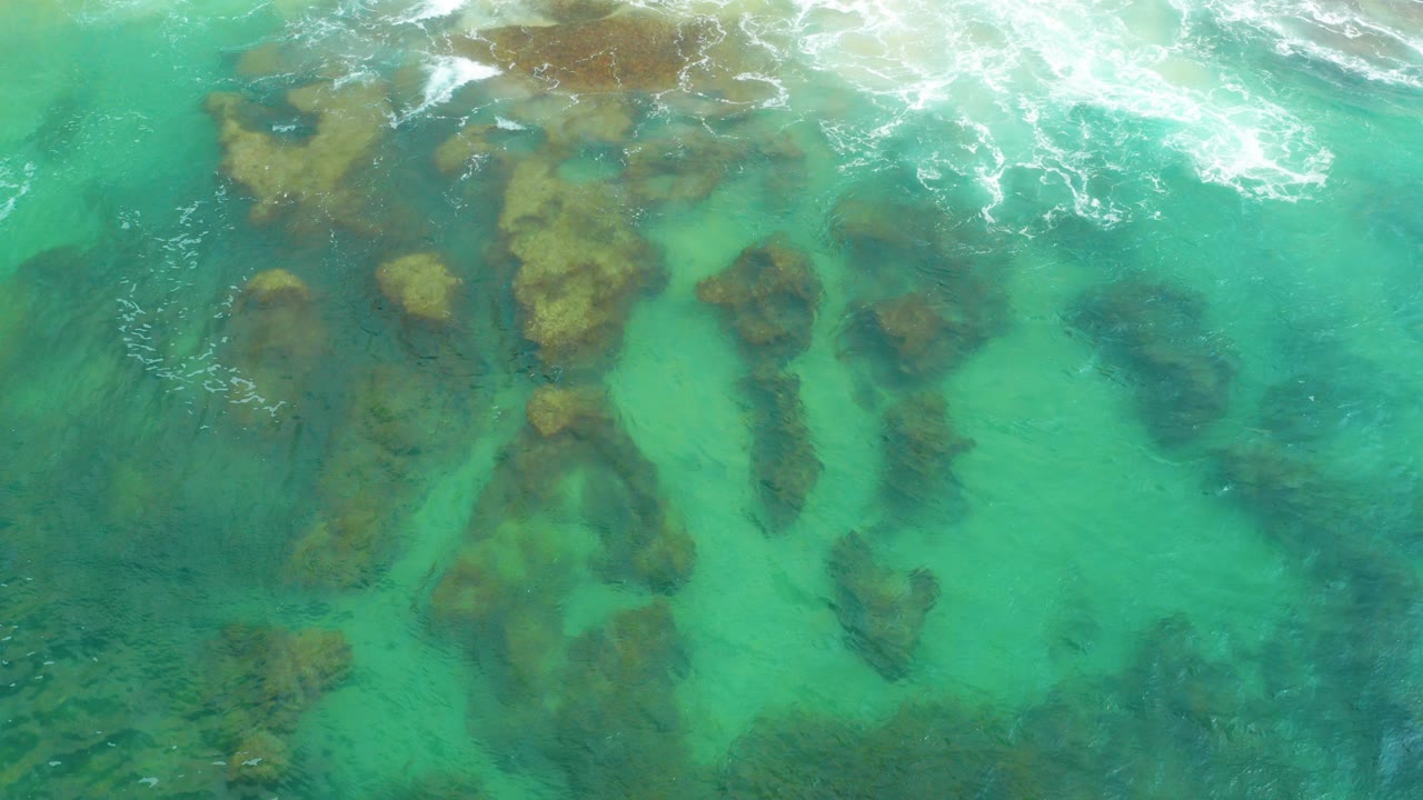 Aerial view over Laguna Beach, Baja California Sur, with turquoise water, dark submerged rocks, white surf along the top edge, and no visible people.