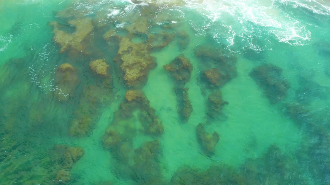 Laguna Beach, Baja California Sur appears from above with foamy wave wash at the top, transparent aquamarine water, scattered dark rocks, and no visible people.