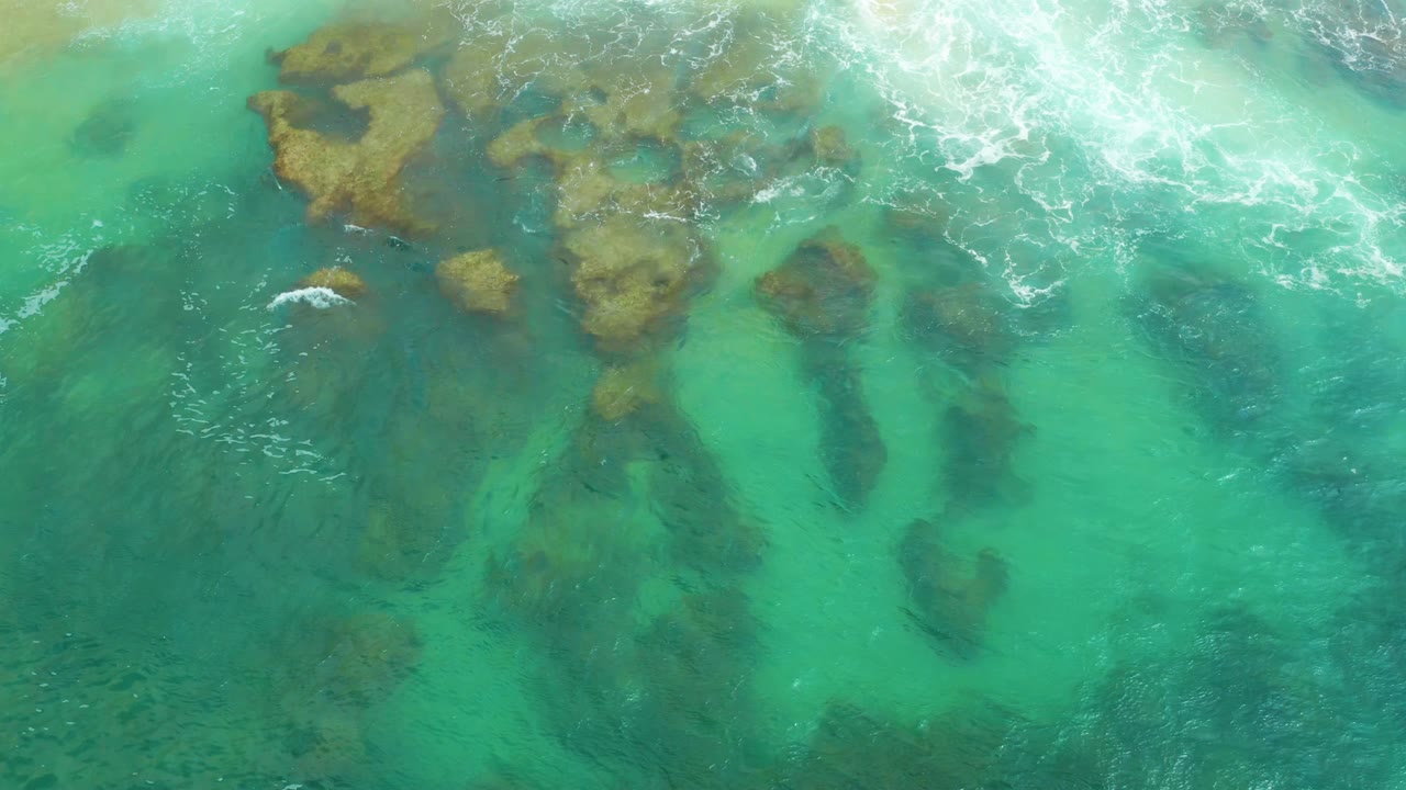 Clear green and turquoise sea covers irregular coral or rock shapes at Laguna Beach, Baja California Sur, with a thin white foam patch near the upper frame and no visible people.