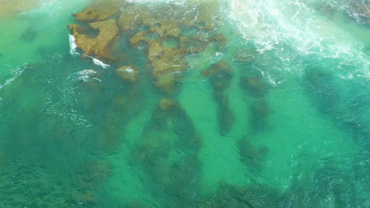 Aerial frame of turquoise ocean over dark and brown reef forms at Laguna Beach, Baja California Sur, with breaking white surf concentrated at the top right and no visible people.