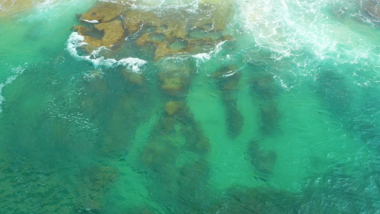 Top-down view of clear turquoise sea and brown submerged rocks at Laguna Beach, Baja California Sur, with white water spreading across the upper band and no visible people.