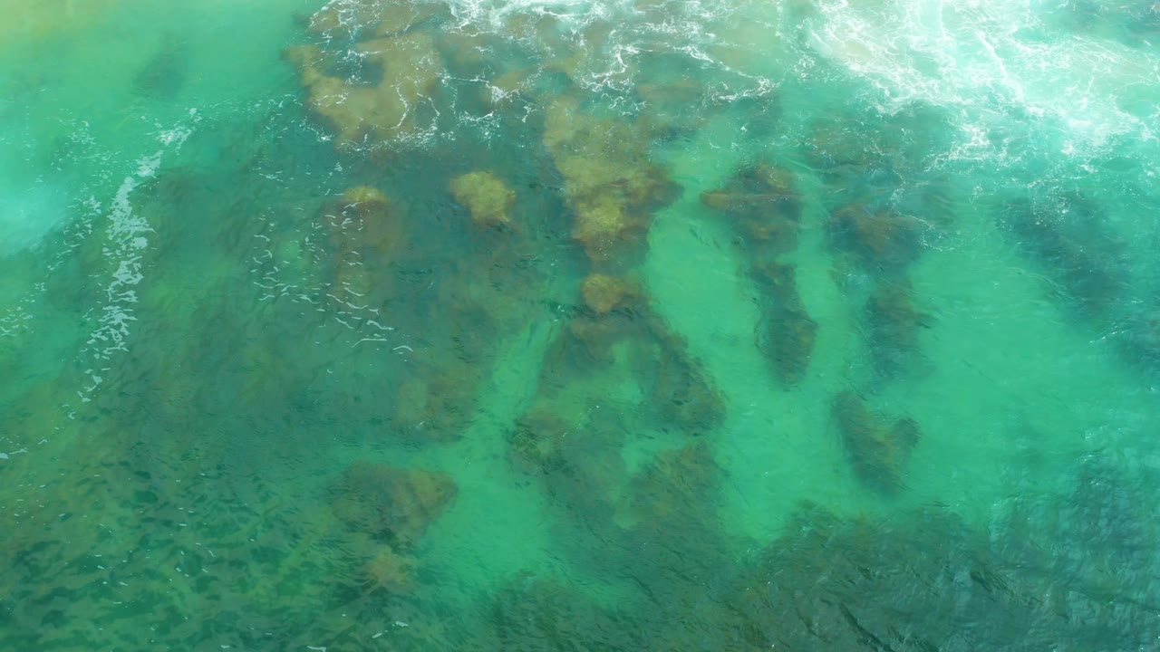 Brown rock shapes sit beneath clear green water in Laguna Beach, Baja California Sur, while a brighter foam band crosses the upper part of the frame and no people are visible.