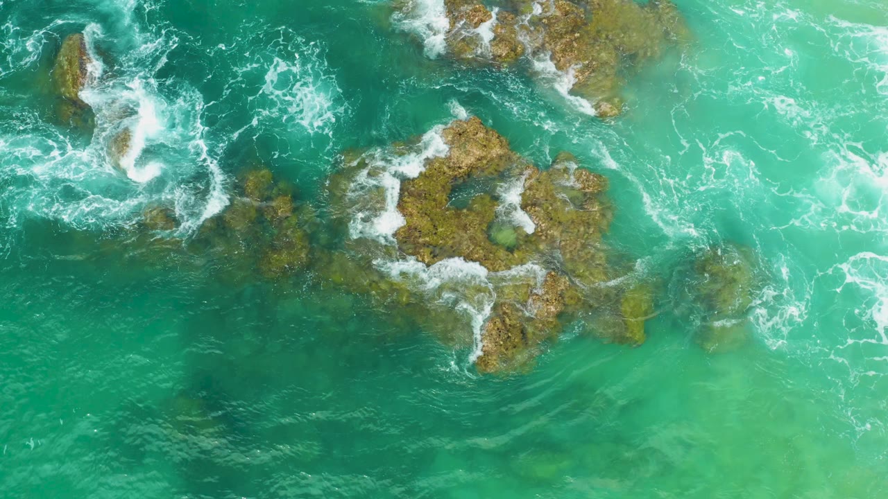 Aerial green ocean shallows show white surf breaking over multiple exposed rocks at Laguna Beach, Baja California Sur, with no visible people.