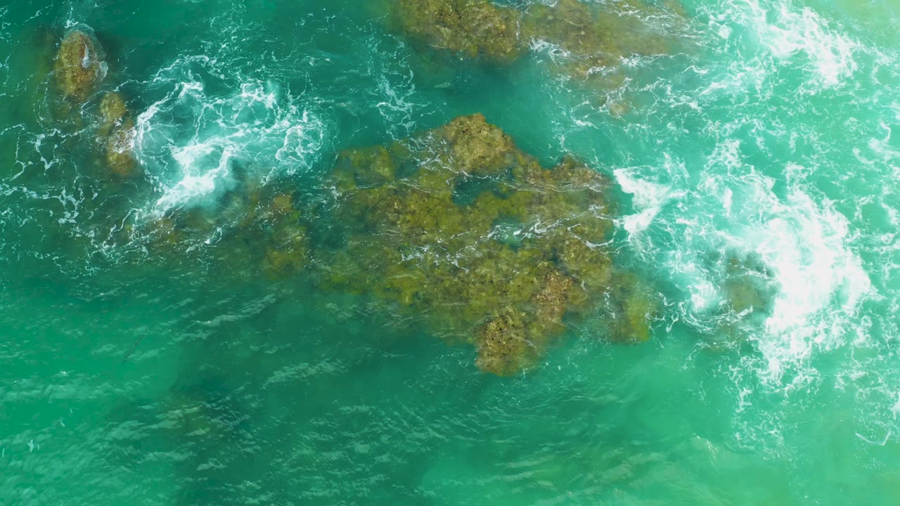 Top-down view of Laguna Beach water where bright foam washes past several coral outcrops and a larger rock patch near center, no visible people.