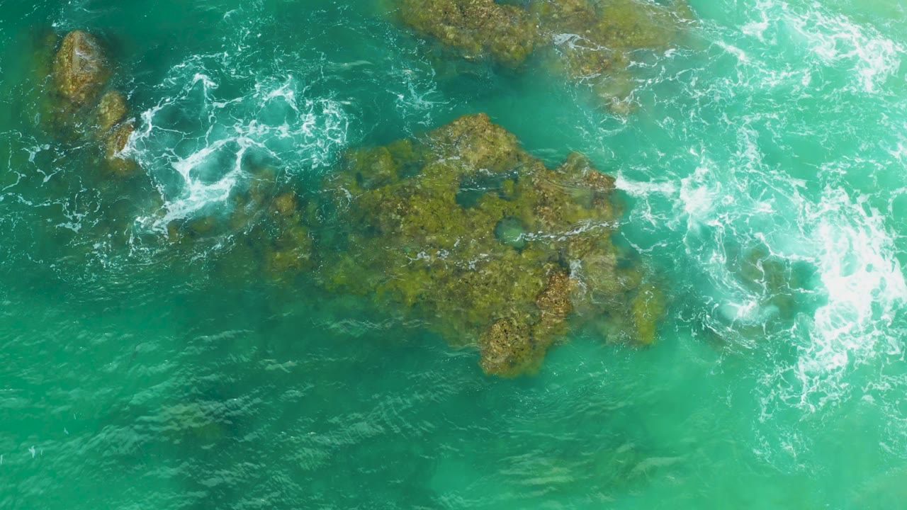 Overhead frame from Laguna Beach with a broad coral rock area under shallow turquoise water and surf pushing in from the right, no visible people.