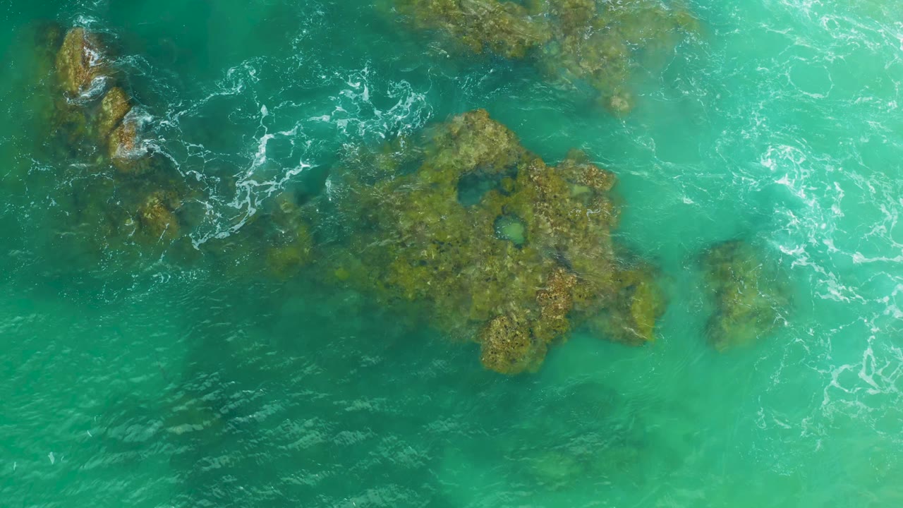 Aerial frame over Laguna Beach reef water, with brown-green rock shapes under turquoise water and thin white foam near the upper-left rocks, no visible people.