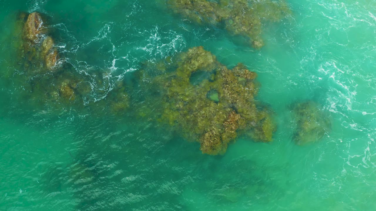 Top-down Laguna Beach ocean frame with reef formations under bright turquoise water, scattered white wave streaks, and no visible people.
