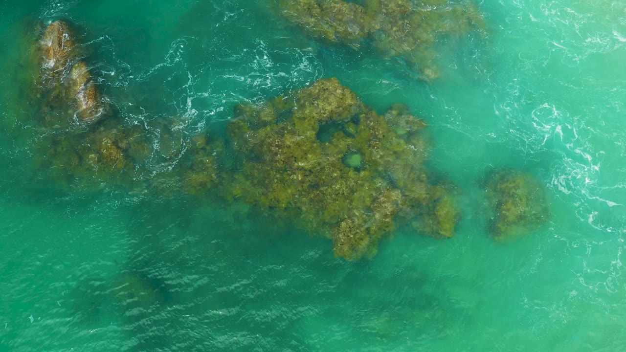 Aerial view of Laguna Beach reef water, with a darker submerged rock area on the left, pale turquoise water on the right, and no visible people.
