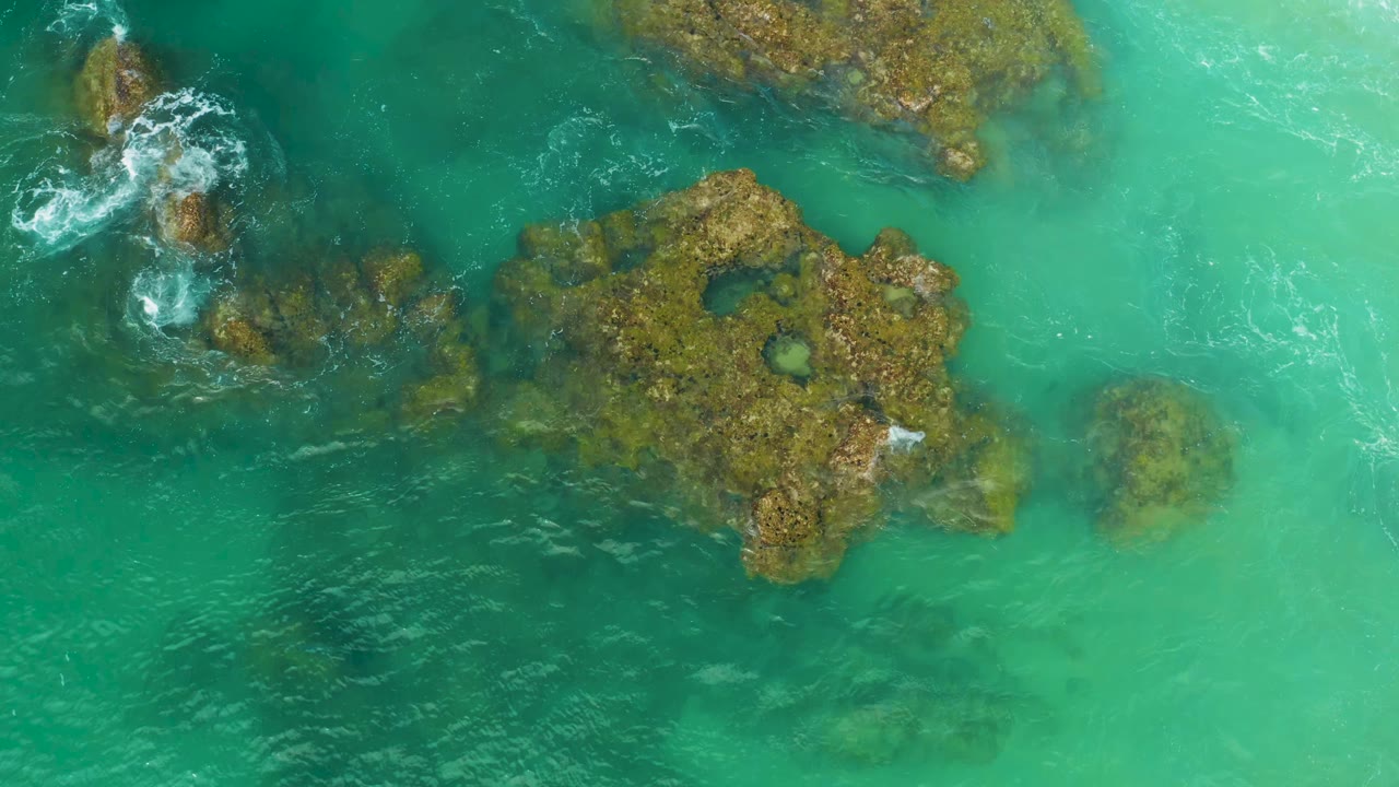 Aerial frame over Laguna Beach, Baja California Sur, with a central brown rock shelf, pale green water to the right, darker water to the left, and no visible people.
