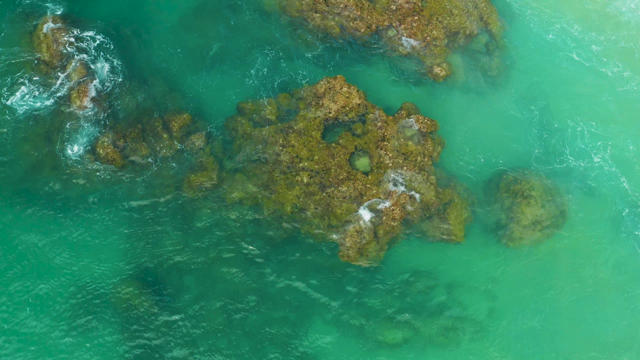 Top-down Laguna Beach, Baja California Sur frame showing rock shelves near the upper and center areas, white foam curling on the left, and no visible people.