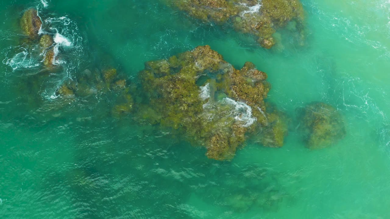 Overhead view at Laguna Beach, Baja California Sur, with a broad central rocky patch, pale clear water at right, foam touching the lower right rocks, and no visible people.