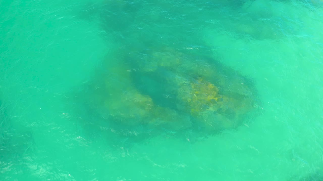 A shallow turquoise water view shows darker rock areas and yellow-brown rock tops beneath the surface in Laguna Beach, Baja California Sur, with no visible people.