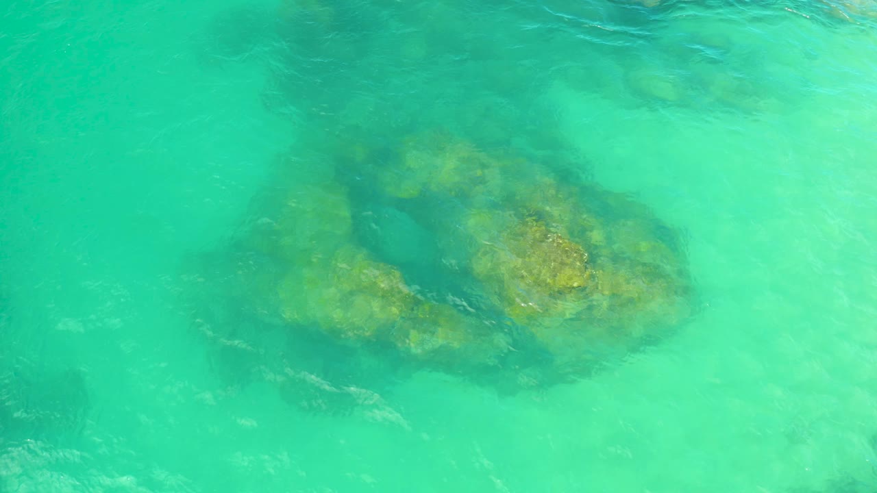 Top-down view of an aqua water surface with a broad submerged rock cluster and darker surrounding patches in Laguna Beach, Baja California Sur, with no visible people.