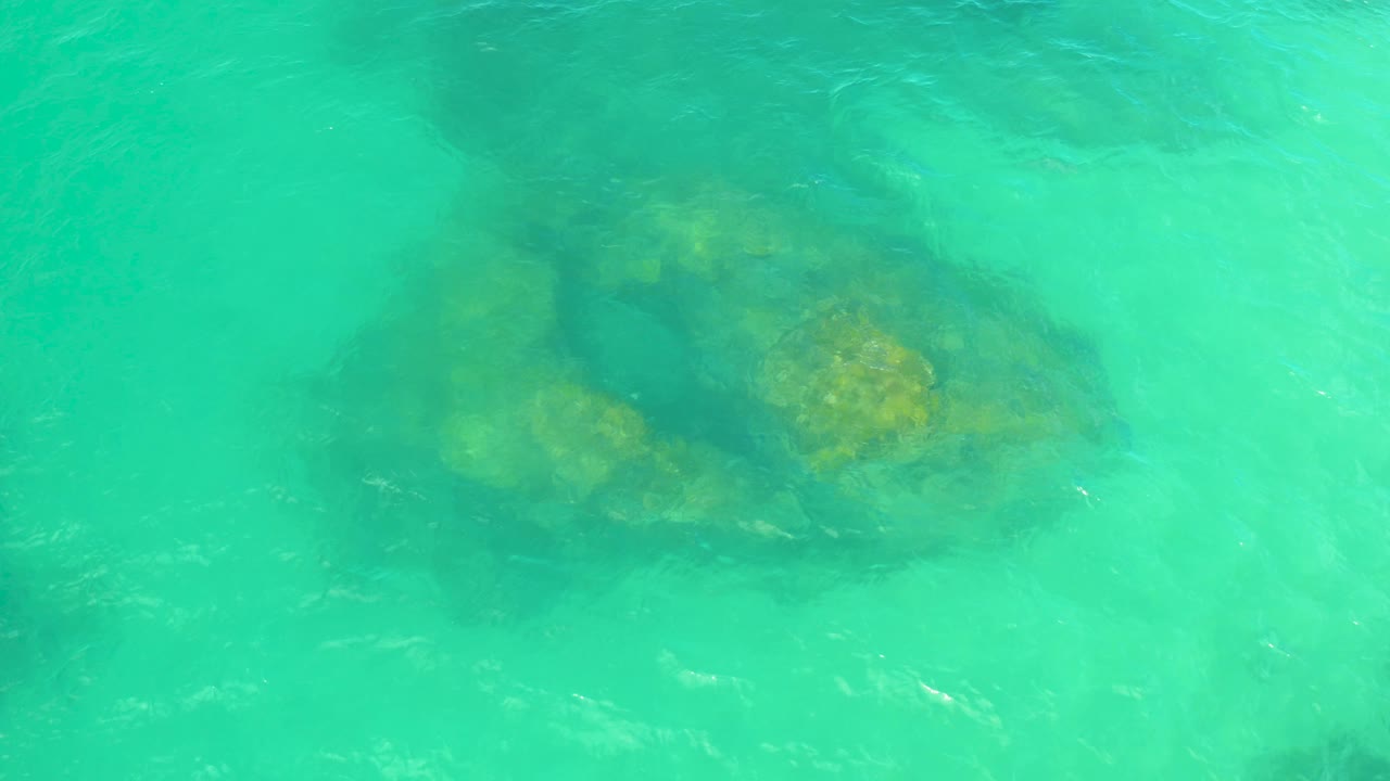 Bright teal shallow ocean water covers a yellow-green rock mass near the upper part of the overhead frame in Laguna Beach, Baja California Sur, with no visible people.