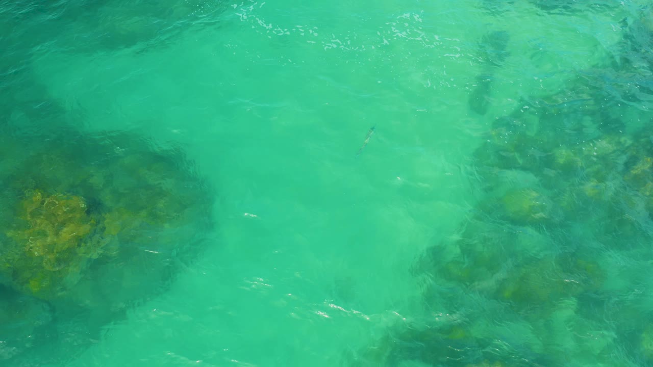 Aerial view of turquoise water in Laguna Beach, Baja California Sur, with darker submerged rocks entering from the left edge and no visible people.
