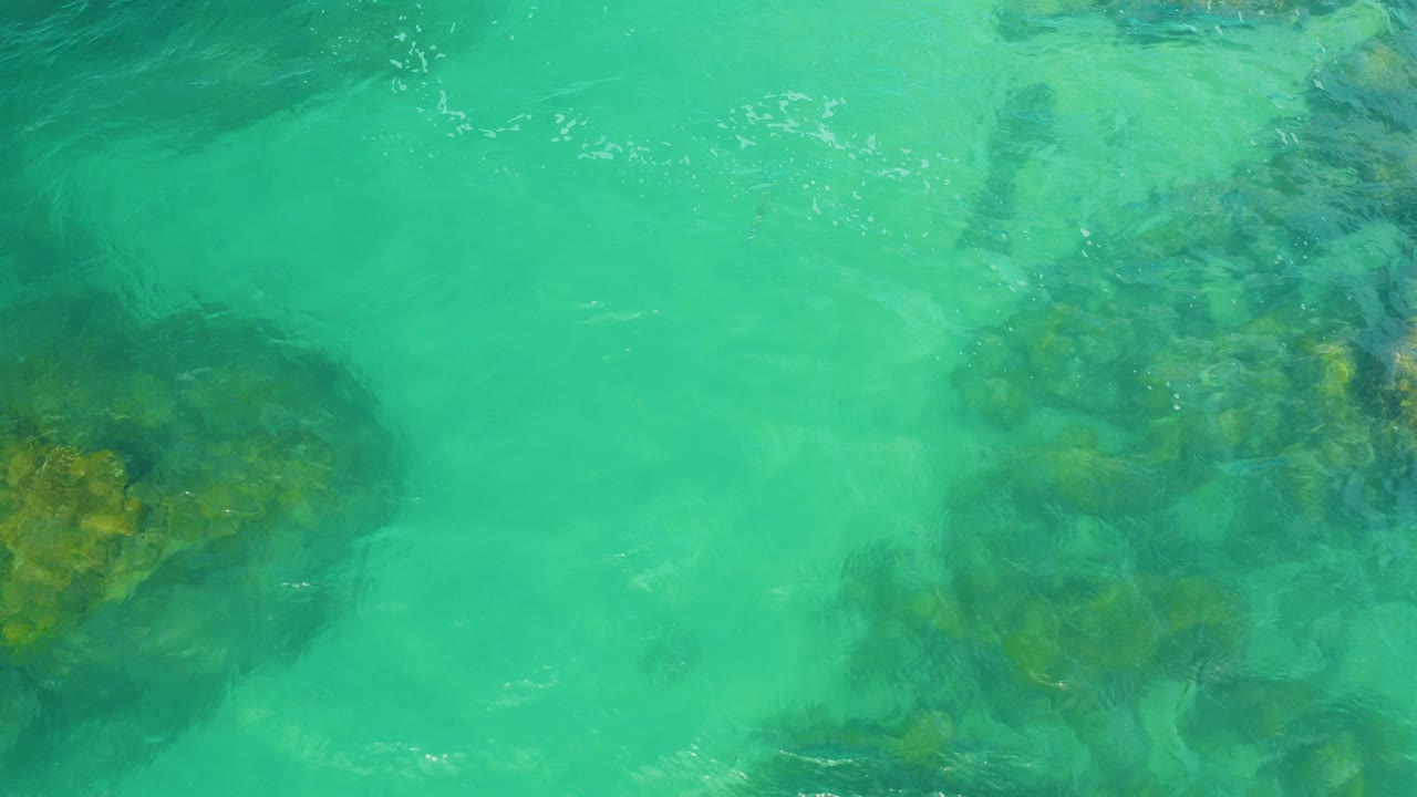 Overhead view of turquoise water in Laguna Beach, Baja California Sur, with submerged rocks at the left and center and no visible people.